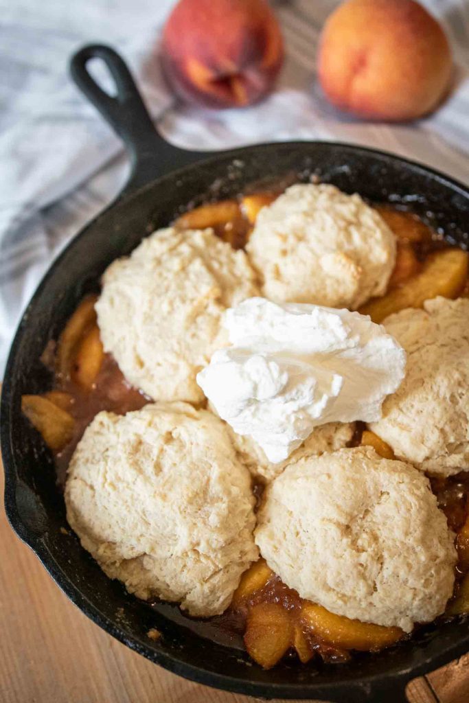 sourdough peach cobbler in a cast iron skillet. fresh peaches lay on a towel in the background