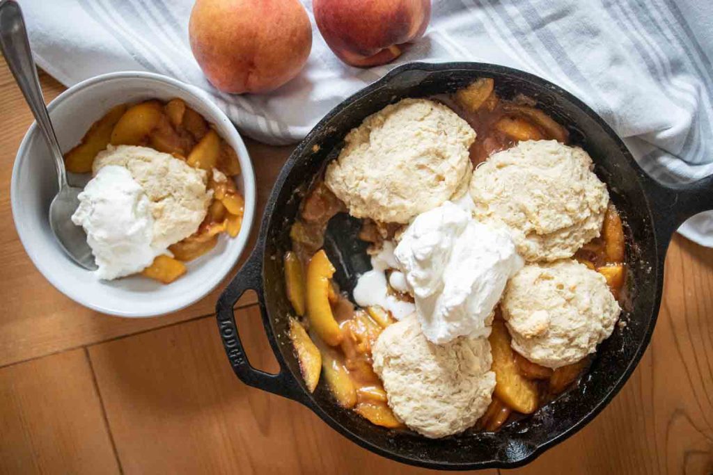 sourdough peach cobbler with single serving in a white bowl with a spoon. fresh peaches lay on a towel in the background
