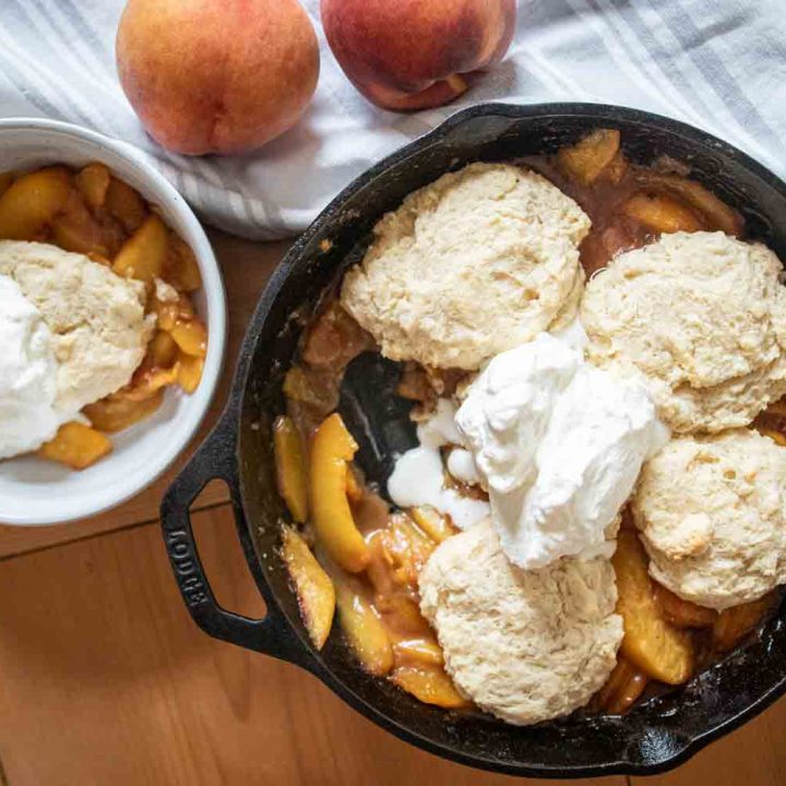 cast iron skillet with sourdough peach cobbler and single serving of peach cobbler in a white bowl