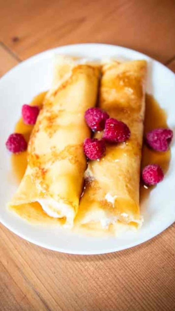 two sourdough crepes on a white plate, stuffed with cream cheese filling and topped with fresh raspberries and maple syrup