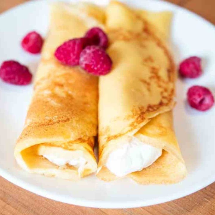 two sourdough crepes on a white plate, stuffed with cream cheese filling and topped with fresh raspberries