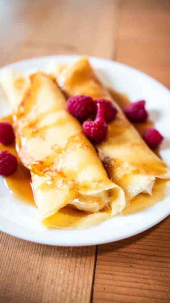 two sourdough crepes on a white plate stuffed with cream cheese filling and topped with fresh raspberries and maple syrup