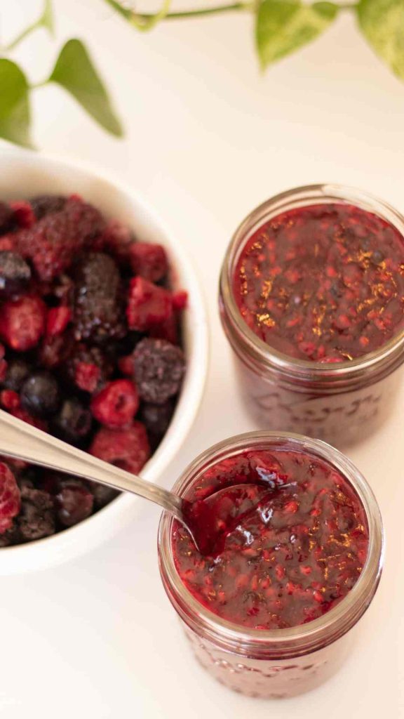 two jars of berry sauce, one with a spoon and a bowl of frozen berries to the left. plant leaves in the background
