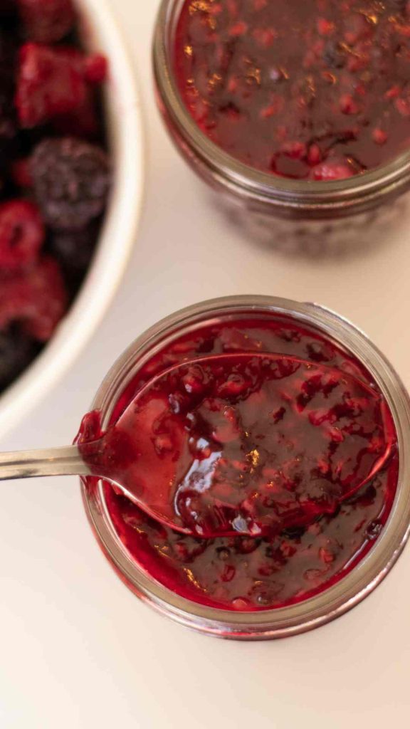 close of up of a spoon holding berry sauce over a full jar. another jar and a bowl of berries in the background