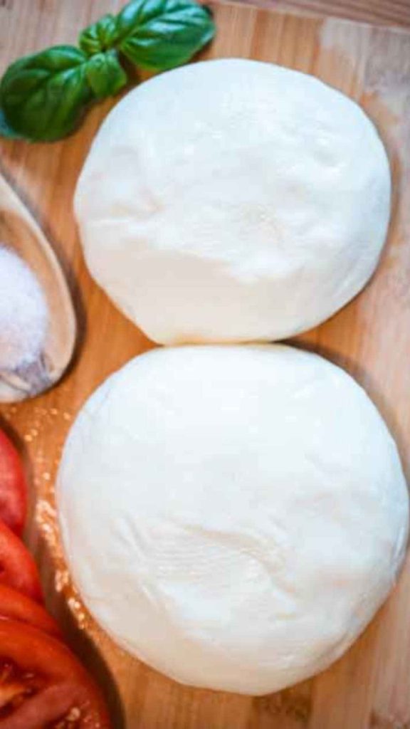 two white balls of raw milk mozzarella cheese surrounded with sliced tomatoes, fresh basil and a wooden spoon of salt