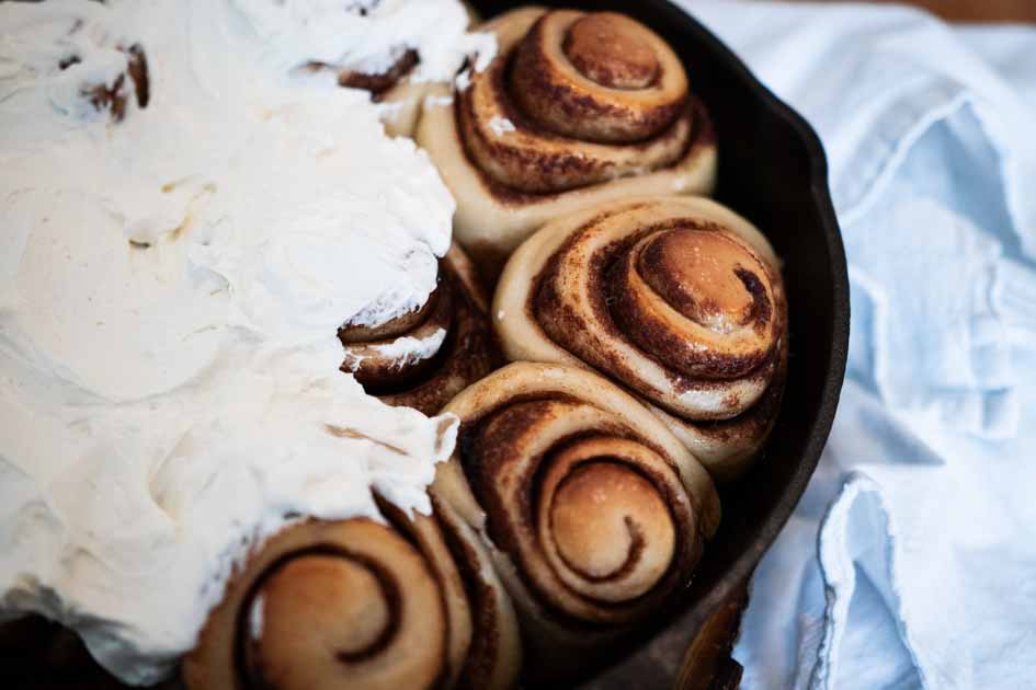 close up of finished sourdough cinnamon rolls in a cast iron skillet with cream cheese frosting on part of it