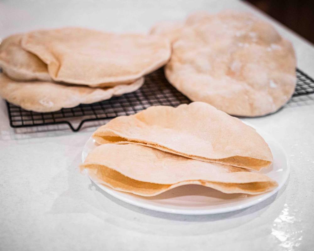 sliced sourdough pita bread on a white plate with pitas on a cooling rack in the background