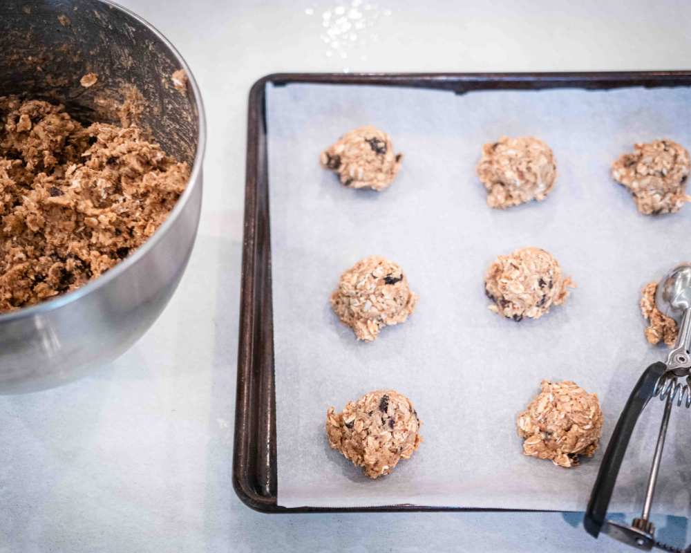 sourdough breakfast cookie dough scooped onto a baking sheet with a cookie scoop. bowl of cookie dough in the background