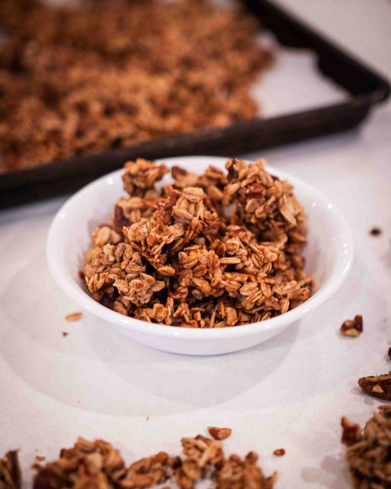 granola in a white bowl with a pan of granola in the background